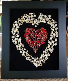 a heart - shaped brooch with pearls and red glass beads is displayed in a black frame