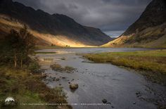 a river running through a lush green valley under a cloudy sky with mountains in the background