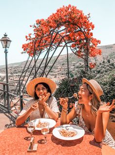 two women sitting at an outdoor table eating pasta and drinking wine, with red flowers in the background