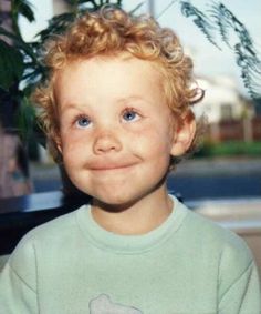 a young boy with curly hair and blue eyes smiling in front of a potted plant