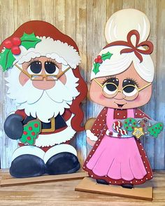 two paper cutouts of santa and mrs claus on wooden planks, one holding a candy cane