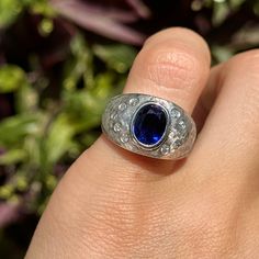 a person's hand holding a ring with a blue stone in it and purple flowers behind it