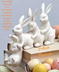 three white ceramic rabbits sitting on top of an open book next to some colorful eggs