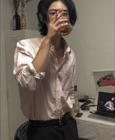 a man drinking from a glass while standing in front of a desk with a laptop