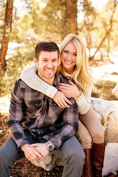 a man and woman sitting on a rock in the woods smiling at the camera with sheep behind them