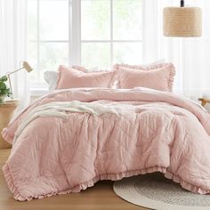 a bed with pink comforter and pillows in front of a window, next to a potted plant