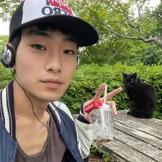 a young man wearing headphones and holding a cup next to a black cat on a park bench