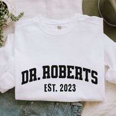 a t - shirt with the words dr roberts est 2023 printed on it next to jeans and a hat