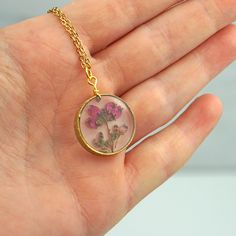 "Real pressed Heather Flowers are encased in jewelry grade resin within this stunning stardust raw brass circle bezel. The setting has a textured finish, which gives just a bit of extra dazzle. Attached to a matching 18\" chain and measures 3/4\" diameter. Hangs on a labelled card, easy for gifting!" Gold Brass Jewelry With Pressed Flowers, Pressed Flowers Yellow Gold Jewelry, Rose Gold Jewelry With Pressed Flowers In Round Pendant, Yellow Gold Jewelry With Pressed Flowers In Round Pendant, Yellow Gold Round Pendant With Pressed Flowers, Botanical Style Gold Resin Jewelry, Resin Round Pendant Jewelry With Birth Flower, Nickel Free Gold Botanical Jewelry, Gold Resin Flower Pendant Jewelry