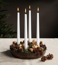 four white candles in a bowl with pine cones and ornaments