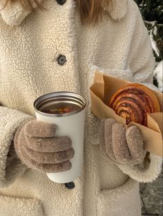 a woman in winter clothing holding a cup of hot chocolate and a donut with mittens