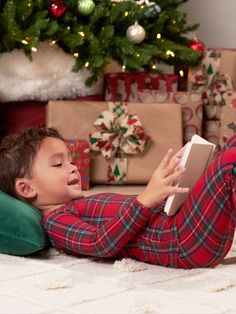 a young boy laying on the floor reading a book in front of a christmas tree