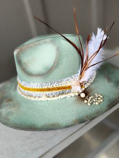 "The perfect sea-foam green hat has been hand painted, and distressed. It's a smaller fedora and has a blue denim paisley band, wrapped in lace and gold velvet. A pearl and gold chain with a pearl tassel is attached. Tied with pink twine is a white feather and brown duck tail feathers.   SIZE: Fedora Hat is OSFA Wired brim measuring 3 3/4\" and a crown height of 4 3/4\". The Fedora Hat has a adjustable band with drawstring which allows for a perfect fit. Materials: This hat is made from a blend of 65% cotton and 35% polyester, providing a comfortable and durable construction. It also includes a faux leather band, suede cord, and with feathers. BENEFITS: - All-Season: This Fedora Hat is suitable for all seasons, allowing you to elevate your style year-round. - Versatile: This wide brim fedo Green Bohemian Hat Bands For Spring, Vintage Fedora Hat For Festivals, Vintage Fedora Mini Hat For Festivals, Green Bohemian Hat Bands For Summer, Bohemian Green Hat Bands For Summer, Bohemian Green Summer Hat Bands, Green Bohemian Hat For Rodeo, Bohemian Green Hats For Rodeo, Vintage Fedora For Festival