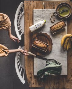 two people sitting at a table with bananas and bread on it, one person holding a knife