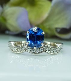 an oval blue sapphire and diamond ring sits on a white surface with flowers in the background