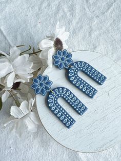 two blue earrings sitting on top of a white plate next to some flowers and leaves