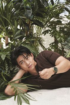 a young man laying on the ground in front of plants and palm trees, wearing a watch