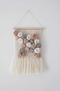 a white wall hanging with flowers and tassels on the bottom, in front of a white wall