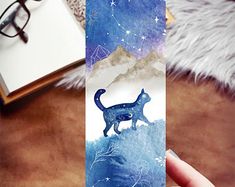 a person holding up a bookmark with a cat on it's side and stars in the sky