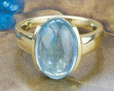 With its subtle sparkle, radiant pale blue tones and simple design, this oval Aquamarine Gemstone Gold Ring exudes sophistication. The smooth, shimmering Aquamarine is polished to reveal its natural tones and inclusions. Framed in a contemporary half setting of Yellow Gold. The natural Aquamarine is centred on a wide band that curves comfortably around your finger. Aquamarine is believed to reduce stress and quiet the mind. DETAILS: Designed and handcrafted in our studio, this ring is a one-off creation. Aquamarine. 7.01 CTS. 9 Ct Yellow Gold setting and band. Measures: Length 15mm X Width 9.5mm. Ring Size: US: 8 (equivalent to UK: P 1/2). This ring can easily be resized if required. Resizing: Resizing takes up to 5 business days. This ring is part of our exclusive Paris Argentina Original Modern Moonstone Ring With Oval Bezel Setting, Oval Modern Moonstone Ring With Bezel Setting, Modern Oval Moonstone Ring With Bezel Setting, Modern Aquamarine Oval Rings, Modern Oval Aquamarine Rings, Modern Oval Blue Topaz Ring, Modern Oval Topaz Ring With Polished Finish, Blue Oval Topaz Ring With Bezel Setting, Modern Blue Topaz Oval Ring