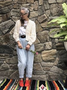 a woman standing in front of a stone wall holding a flower and wearing bright colored shoes