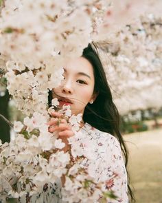 a woman standing under a tree with white flowers on it's branches and looking at the camera