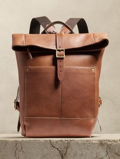 a brown leather backpack sitting on top of a stone slab with the straps down and two buckles at the bottom