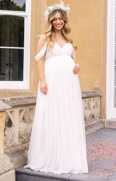 a pregnant woman wearing a white dress standing in front of a building with flowers on her head