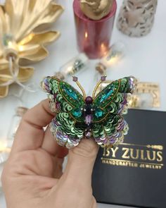 a hand holding a colorful butterfly brooch in front of a box with other items