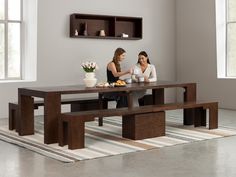 two women sitting at a wooden table with plates of food in front of them on a rug