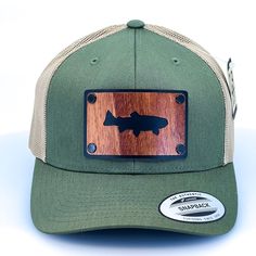 Introducing our stylish and durable olive green and khaki trucker cap, perfect for any outdoor adventure. The hat features a special patch made exclusively in our shop in Missoula, Montana. The patch is made of high-quality mahogany veneer and has been cut out in the shape of a trout, laminated to a black anodized aluminum plate, and secured with black rivets. The design on the mahogany veneer adds a unique touch to the hat with its intricate details and natural wood grain. The cap features a br Brown Military Style Baseball Cap For Outdoor, Brown Military Baseball Cap For Outdoor, Brown Military Style Trucker Hat, Brown Military Trucker Hat, Khaki Curved Bill Snapback Hat For Outdoor, Green Snapback Hat With Logo Patch For Outdoor, Khaki Cap For Fishing, Khaki Fishing Cap, Khaki Snapback Trucker Hat For Outdoor Activities