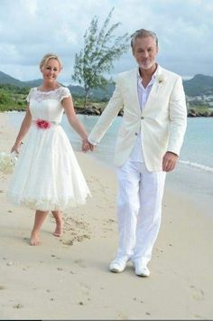 a man and woman are walking on the beach holding hands while dressed in wedding attire