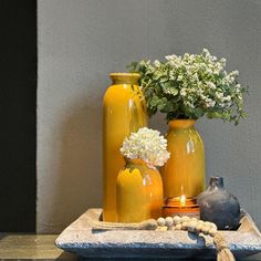 three yellow vases with white flowers in them on a tray next to a gray wall