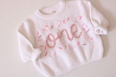 Beautiful hand embroidered ONE chunky sweater. Perfect for your baby's first birthday.  This listing is for one sweater only. You pick the wording from 1/2, ONE, or TWO White Sweater and pink lettering plus sprinkles as pictured. If you want a different color, just send us a convo. We'll be happy to customize for you Hand embroidery on a chunky sweater True to size Custom orders welcome Cute White Sweater With Embroidered Text, White Cute Sweater With Embroidered Text, Cute Crew Neck Birthday Sweater, Cute Crew Neck Sweater For Birthday, Cute Sweater For Birthday In Winter, Cotton Crew Neck Sweater For Birthday, Cute Cotton Sweater For Birthday, Embroidered Winter Birthday Sweater, Hand Embroidered Sweater