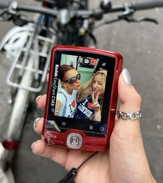 a person holding up a red cell phone with two girls on it and a bike in the background