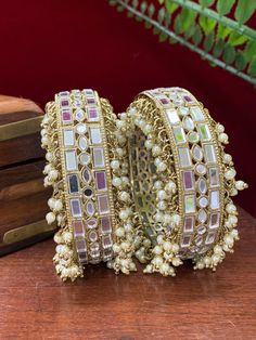 This authentic Kalinda bangle is a classic blend of traditional aesthetics and modern craftsmanship. Intricately crafted of brass with real mirror and pearl work, its one-inch width and champagne stone sparkle make it a chic, timeless addition to any ensemble. Comes in a pair. Elegant Kundan Bangle For Festive Occasions, Elegant Gota Work Bangle As Gift, Elegant Gota Work Bangle For Gifts, Gold Bohemian Pearl Bracelet For Wedding, Elegant Kundan Bangle With Stone Work, Party Bracelets In Kundan With Gota Work, Elegant Bangle With Gota Work For Festivals, White Temple Jewelry Bangle With Stone Work, White Stone Work Temple Jewelry Bangle