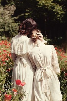 two women in white dresses are walking through the field with red poppies and trees behind them