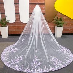 a white veil with purple flowers on the bottom is hanging in front of a brick wall