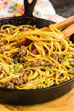 a skillet filled with noodles and meat on top of a wooden table next to two spoons