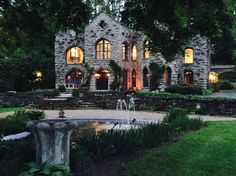 a large stone house with a fountain in the front yard at night, surrounded by greenery and trees