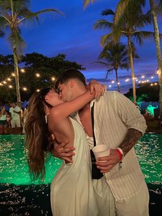 a man and woman are kissing in front of a pool at night with palm trees