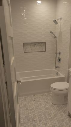 a white toilet sitting next to a bath tub under a window in a bathroom with hexagonal tiles on the floor