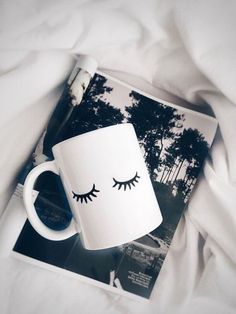 a coffee mug with eyelashes on it sitting on top of a white bed sheet next to a magazine