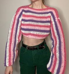 a woman wearing a pink and purple crochet top with long sleeves, standing in front of a white wall