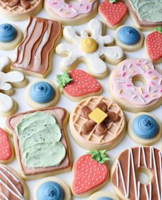 many decorated cookies and pastries on a white tablecloth with blue, pink, green, orange, and yellow icing