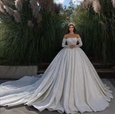 a woman in a white wedding dress standing next to some tall grass and plants with her hands on her hips