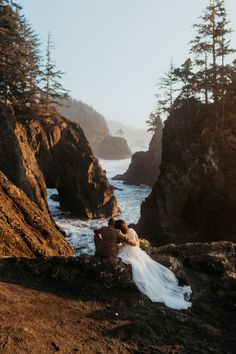 a bride and groom are sitting on the rocks by the ocean in front of some trees