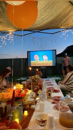 a group of people sitting at a long table with food and drinks in front of a screen