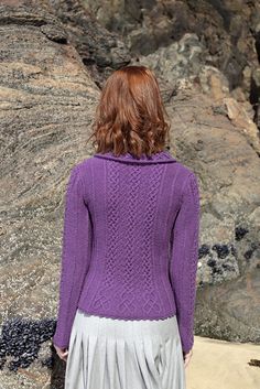 a woman wearing a purple sweater and pleated skirt standing in front of some rocks