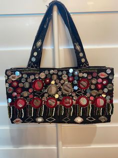 This Moyna handbag is a unique and stylish accessory. With its colorful exterior and intricate beaded and mirrored accents all around, it is sure to turn heads. The bag is small, measuring 10 inches in width, 5 inches in height, and 3 inches in depth, but it is perfect for carrying all your essentials and has a small interior pocket. The bag features a snap closure and a multicolor handle with a 6-inch drop. The interior is lined with black satin. The lightweight and packable design makes it per Beaded Tote Bag For Shopping, Multicolor Evening Bags For Festivals, Festive Beaded Handheld Bags, Multicolor Embellished Bags For Festive Occasions, Embellished Pouch Shoulder Bag For Festivals, Beaded Shoulder Bag As Fashion Accessory, Multicolor Beaded Handheld Shoulder Bag, Festive Beaded Pouch Shoulder Bag, Beaded Pouch Shoulder Bag For Festive Occasions